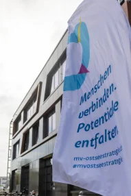 Eine weiße Fahne mit dem Logo und Slogan der „MV-Ostseestrategie“ („Menschen verbinden. Potentiale entfalten.“) weht im Vordergrund vor der modernen Außenfassade des Veranstaltungsgebäudes.