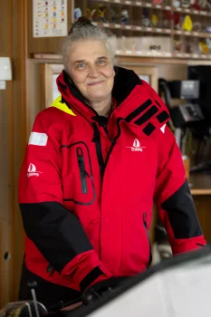 Dr. Regine Labrenz mit grauen, hochgesteckten Haaren trägt eine markante rote Segel- oder Schlechtwetterjacke mit schwarzen Einsätzen. Sie steht im Inneren eines Schiffes.