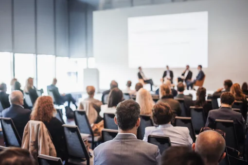 Blick von hinten über die Schultern des Publikums auf eine Podiumsdiskussion in einem großen, hellen Konferenzsaal. Auf der Bühne sitzt eine vierköpfige Diskussionsrunde vor einer großen, weißen Projektionsfläche.
