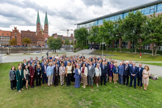 Gruppenbild der 35. Ostseeparlamentarierkonferenz in Lübeck Teilnehmende der 35. Ostseeparlamentarierkonferenz stehen in mehreren Reihen auf einer Rasenfläche in Lübeck und blicken in die Kamera. Im Hintergrund sind die Trave, historische Backsteinhäuser und zwei hohe Kirchtürme zu sehen.