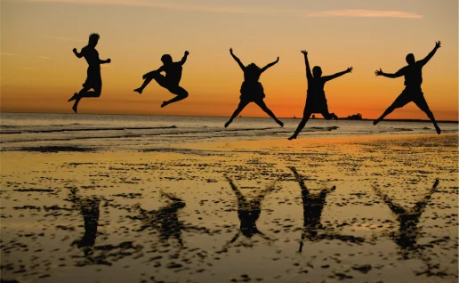 Jugendliche springen am Strand bei Sonnenuntergang Silhouetten von fünf jungen Menschen, die bei Sonnenuntergang am Strand in die Luft springen, mit Spiegelungen im nassen Sand.