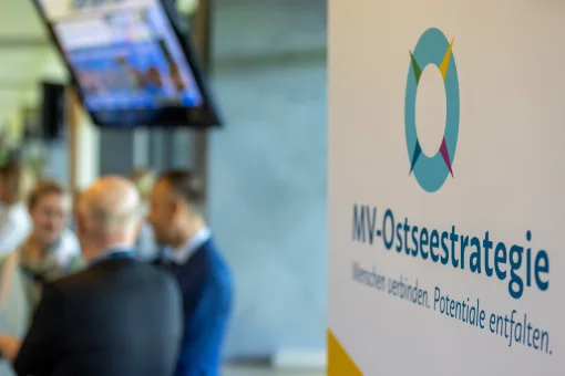 Nahaufnahme eines Informationsbanners mit dem Logo und Slogan der „MV-Ostseestrategie“. Im unscharfen Hintergrund sind Teilnehmende einer Veranstaltung der MV-Ostseekooperation im Gespräch zu sehen.