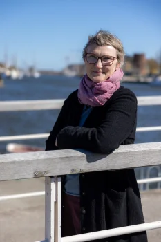 Porträt von Dr. Gudrun Mernitz (Witeno GmbH). Sie trägt Brille, schwarzen Mantel und rosa Schal und lehnt an einem Holzgeländer. Im unscharfen Hintergrund ist ein sonniger Hafen zu sehen.