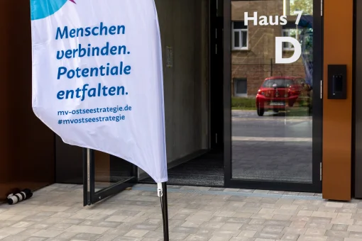 Eine weiße Beachflag mit dem Logo und Slogan der „MV-Ostseestrategie“ („Menschen verbinden. Potentiale entfalten.“) steht vor dem Eingang von „Haus 7 D“ des modernen Veranstaltungsgebäudes.