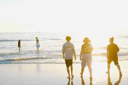 Fünf junge Menschen stehen und gehen bei tief stehender Sonne am Strand. Drei von ihnen bewegen sich barfuß auf das Meer zu, während zwei bereits im flachen Wasser stehen. Das warme Gegenlicht spiegelt sich auf den Wellen und dem nassen Sand.