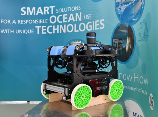 Ein moderner Unterwasser-Erkundungsroboter von Fraunhofer mit schwarzem Rahmen, blauen Elementen und vier auffälligen, hellgrünen Rädern in Wabenoptik steht auf einem Tisch. Im Hintergrund befindet sich eine blaue Messewand.