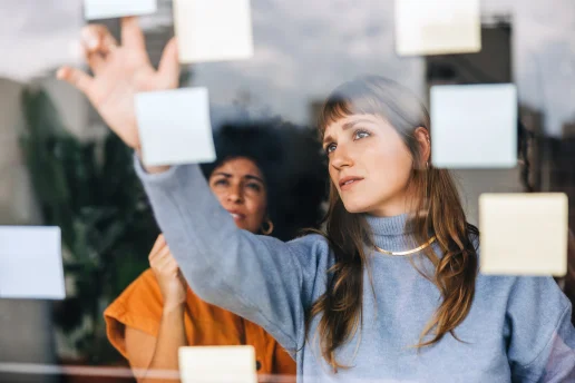 Zwei Frauen arbeiten konzentriert während einer Brainstorming-Sitzung zusammen. Eine Frau im hellblauen Rollkragenpullover bringt Haftnotizen an einer Glasscheibe an, während ihre Kollegin im Hintergrund aufmerksam zuschaut.