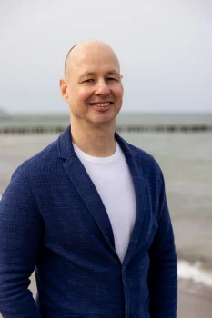 Ein Porträt von Lars Bauer (BioCon Valley) im blauen Sakko, der lächelnd am Ostseestrand steht. Im Hintergrund ist das Meer mit Buhnen zu sehen.
