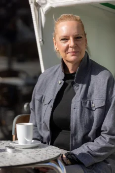 Porträt von Mandy Kröppelien (EIZ Rostock). Sie sitzt an einem sonnigen Tag draußen an einem kleinen Tisch mit einer weißen Kaffeetasse und trägt eine graue Jacke.
