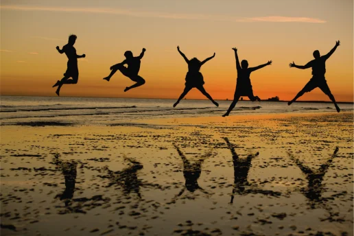 Silhouetten von fünf jungen Menschen, die bei Sonnenuntergang am Strand in die Luft springen, mit Spiegelungen im nassen Sand.