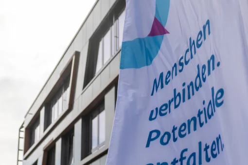 Eine weiße Fahne mit dem Logo und Slogan der „MV-Ostseestrategie“ („Menschen verbinden. Potentiale entfalten.“) weht im Vordergrund vor der modernen Außenfassade des Veranstaltungsgebäudes.