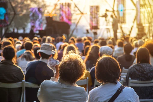 Blick von hinten auf ein großes Publikum, das in Stuhlreihen ein Open-Air-Konzert besucht. Die Szene ist in das warme Licht der tiefstehenden Abendsonne getaucht, während im Hintergrund eine Bühne mit einem Flügel zu erkennen ist.
