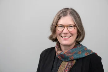 Porträt von Ruth Slenczka. Sie blickt lächelnd in die Kamera, trägt eine Brille, eine dunkle Jacke und einen bunt gemusterten Schal vor einem schlichten, grauen Hintergrund.
