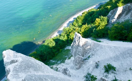 Blick von oben auf die markanten weißen Kreidefelsen auf der Insel Rügen. Die steilen Klippen fallen hinab zu einem schmalen Strand am türkisblauen Wasser der Ostsee. Die Hänge sind teilweise mit dichtem, grünem Wald bewachsen.