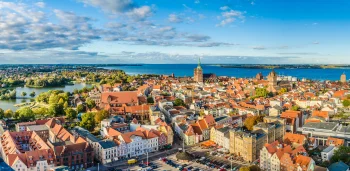 Ein Panorama-Blick von oben auf die historische Altstadt von Stralsund mit ihren charakteristischen roten Hausdächern, den großen Backsteinkirchen St. Jakobi und St. Nikolai sowie dem Knieperteich auf der linken Seite. Im Hintergrund ist die Ostsee.
