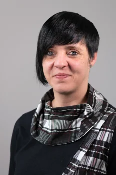 Porträt von Franziska Jende. Sie blickt freundlich in die Kamera und trägt ein dunkles Oberteil mit einem auffällig karierten Kragen. Der Hintergrund ist in schlichtem Grau gehalten.