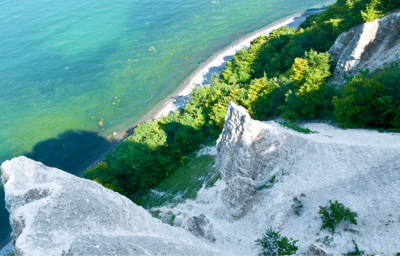 Blick von oben auf die markanten weißen Kreidefelsen auf der Insel Rügen. Die steilen Klippen fallen hinab zu einem schmalen Strand am türkisblauen Wasser der Ostsee. Die Hänge sind teilweise mit dichtem, grünem Wald bewachsen.