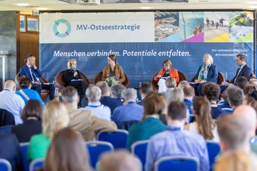 Eine Podiumsdiskussion auf einer Konferenz der MV-Ostseekooperation. Sechs Teilnehmende sitzen in Sesseln auf der Bühne vor einem großen Hintergrundbanner mit der Aufschrift „MV-Ostseestrategie“, während das Publikum im Vordergrund zuhört.
