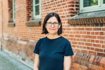 Porträt von Margit Bussmann. Sie trägt eine Brille sowie ein dunkelblaues T-Shirt und steht lächelnd vor einer Wand aus roten Backsteinen.