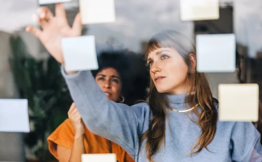 Zwei Frauen arbeiten konzentriert während einer Brainstorming-Sitzung zusammen. Eine Frau im hellblauen Rollkragenpullover bringt Haftnotizen an einer Glasscheibe an, während ihre Kollegin im Hintergrund aufmerksam zuschaut.
