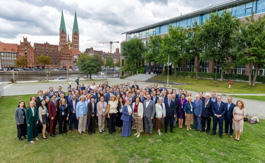 Gruppenbild der 35. Ostseeparlamentarierkonferenz in Lübeck Teilnehmende der 35. Ostseeparlamentarierkonferenz stehen in mehreren Reihen auf einer Rasenfläche in Lübeck und blicken in die Kamera. Im Hintergrund sind die Trave, historische Backsteinhäuser und zwei hohe Kirchtürme zu sehen.
