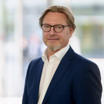 Porträt von Andree Iffländer (Windenergy Network e.V.). Er trägt eine Brille, ein blaues Sakko sowie ein weißes Hemd und blickt lächelnd vor einem hellen, unscharfen Hintergrund in die Kamera.