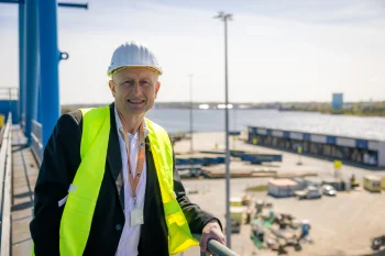 Porträt von Dr. Gernot Tesch (Rostock Port) mit Schutzhelm und gelber Warnweste. Er steht lächelnd auf einer Plattform mit Blick über das Hafengelände.