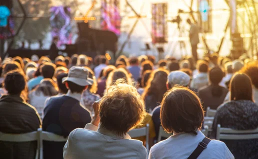 Blick von hinten auf ein großes Publikum, das in Stuhlreihen ein Open-Air-Konzert besucht. Die Szene ist in das warme Licht der tiefstehenden Abendsonne getaucht, während im Hintergrund eine Bühne mit einem Flügel zu erkennen ist.