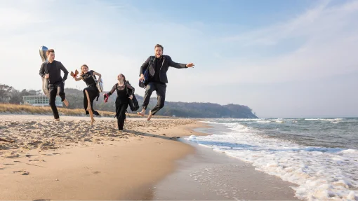 MV-Ostseekooperation - Musiker springen am Strand Vier Personen in eleganter, dunkler Kleidung springen barfuß und fröhlich am Strand der Ostsee in die Luft. Sie tragen Instrumentenkoffer auf dem Rücken. Im Hintergrund sind sanfte Wellen, ein Strandabschnitt und ein bewaldeter Küstenstreifen zu sehen.
