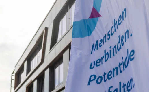 Eine weiße Fahne mit dem Logo und Slogan der „MV-Ostseestrategie“ („Menschen verbinden. Potentiale entfalten.“) weht im Vordergrund vor der modernen Außenfassade des Veranstaltungsgebäudes.