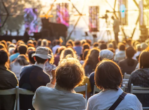 Blick von hinten auf ein großes Publikum, das in Stuhlreihen ein Open-Air-Konzert besucht. Die Szene ist in das warme Licht der tiefstehenden Abendsonne getaucht, während im Hintergrund eine Bühne mit einem Flügel zu erkennen ist.